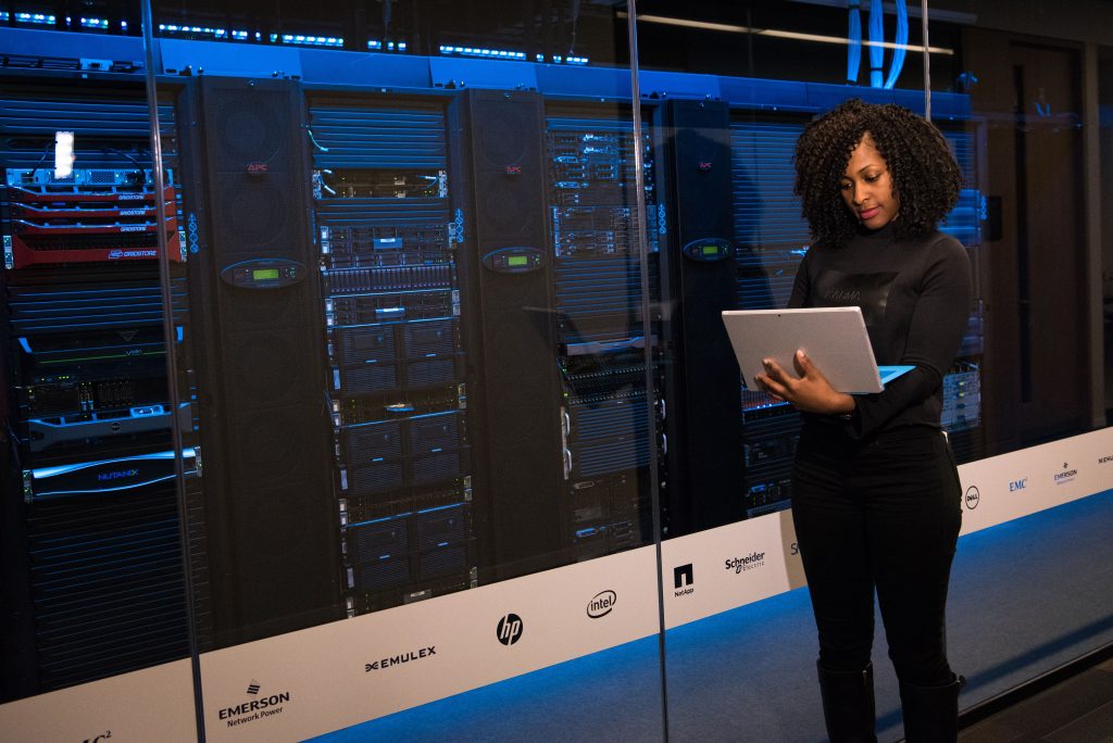 Services A female engineer using a laptop while monitoring data servers in a modern server room.