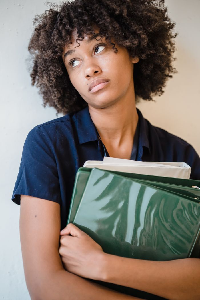 Young woman with curly hair holding files, looking thoughtful indoors. Perfect for business and office concepts.